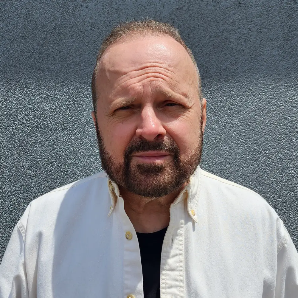 Close-up portrait of a middle-aged man with a beard, wearing a light button-down shirt against a gray textured background.